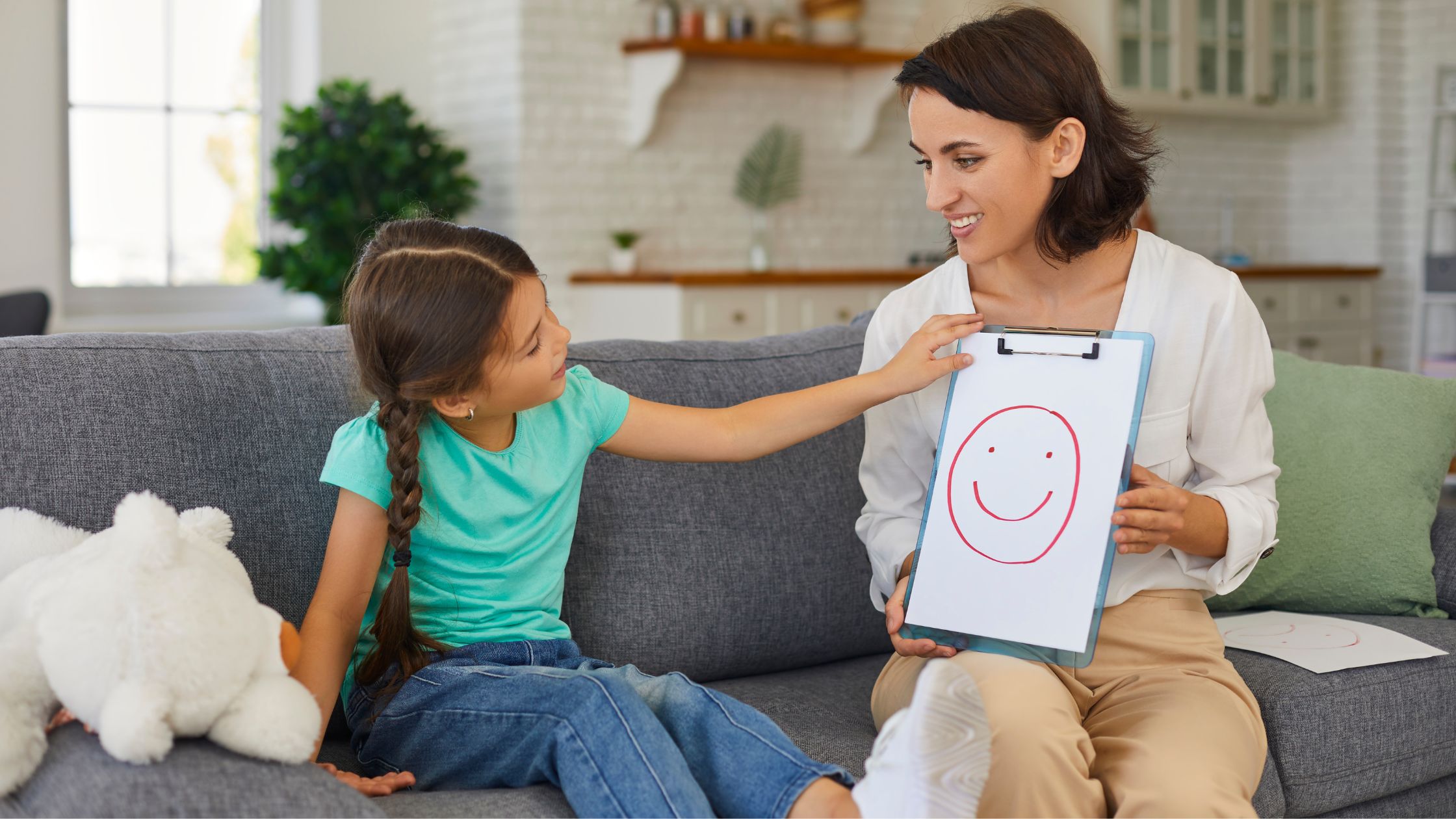 Smiling therapist holding a clipboard with a hand-drawn smiley face as a young girl excitedly points at it, highlighting creative expression in art therapy.