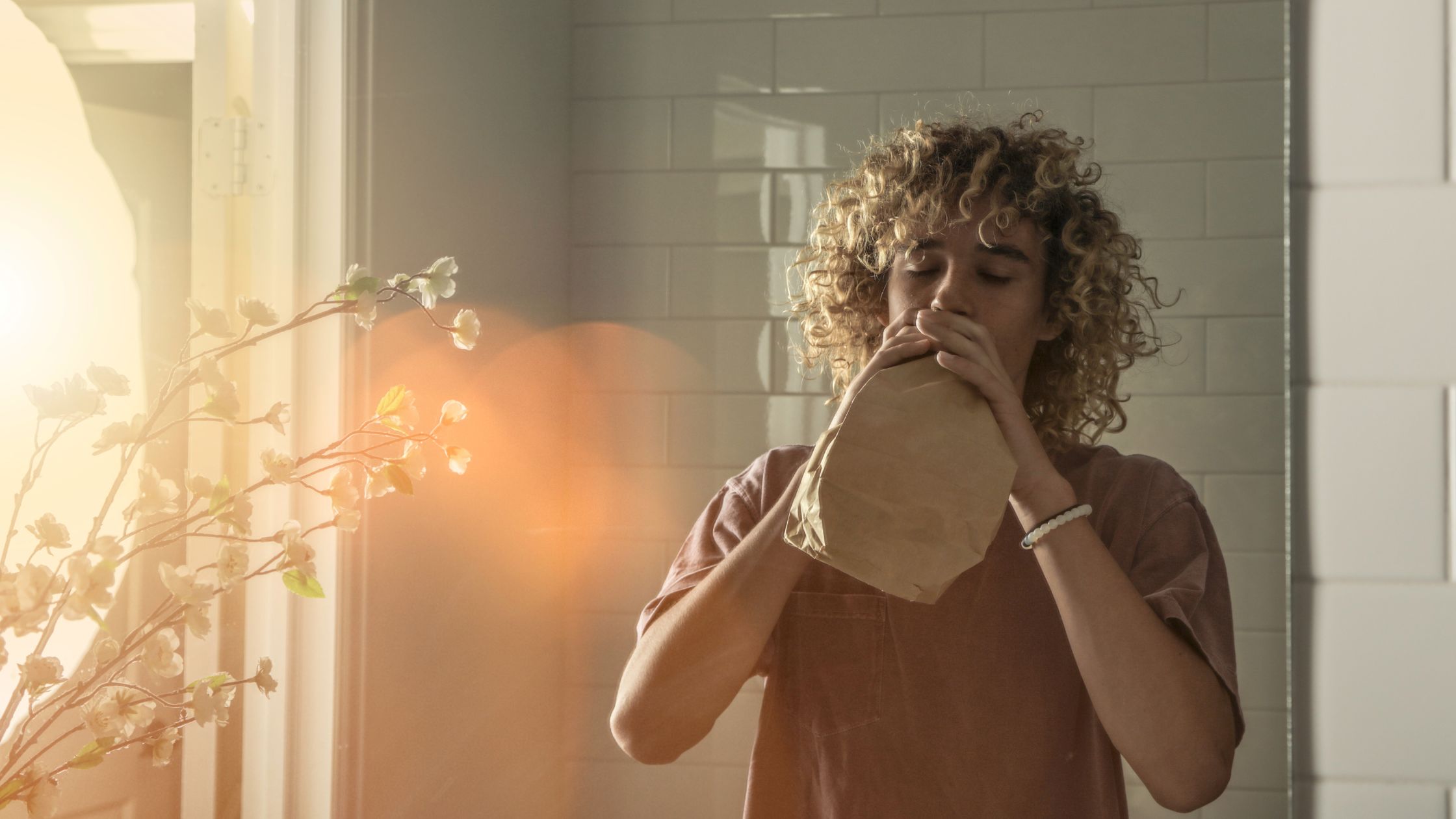 Teen with curly hair using a paper bag to regulate breathing in a sunlit room. Holistic teen treatment approaches at Nexus Teen Academy for emotional wellness.