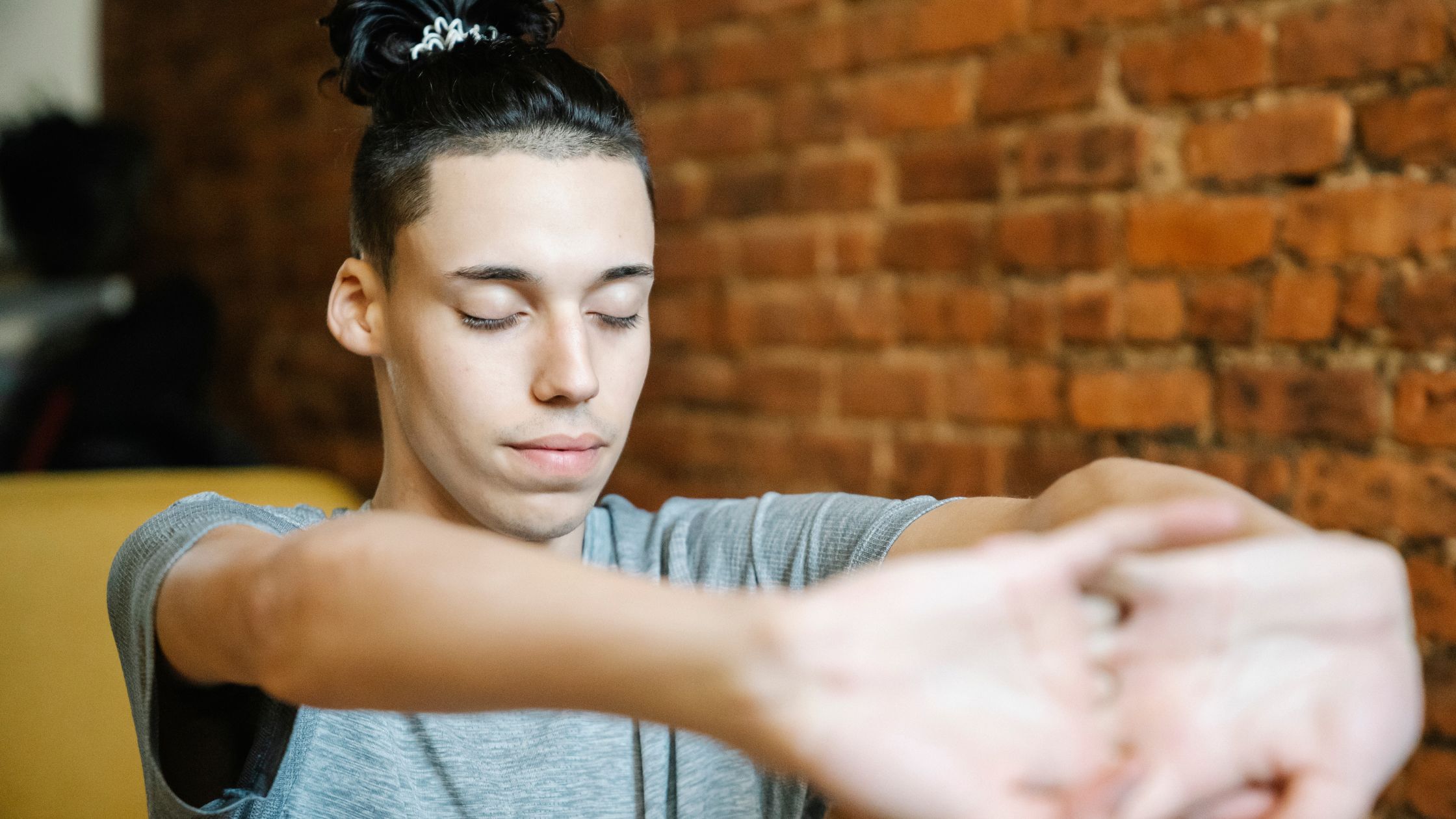 Teen practicing meditation with eyes closed and arms stretched, sitting in a cozy indoor space with a brick wall background.