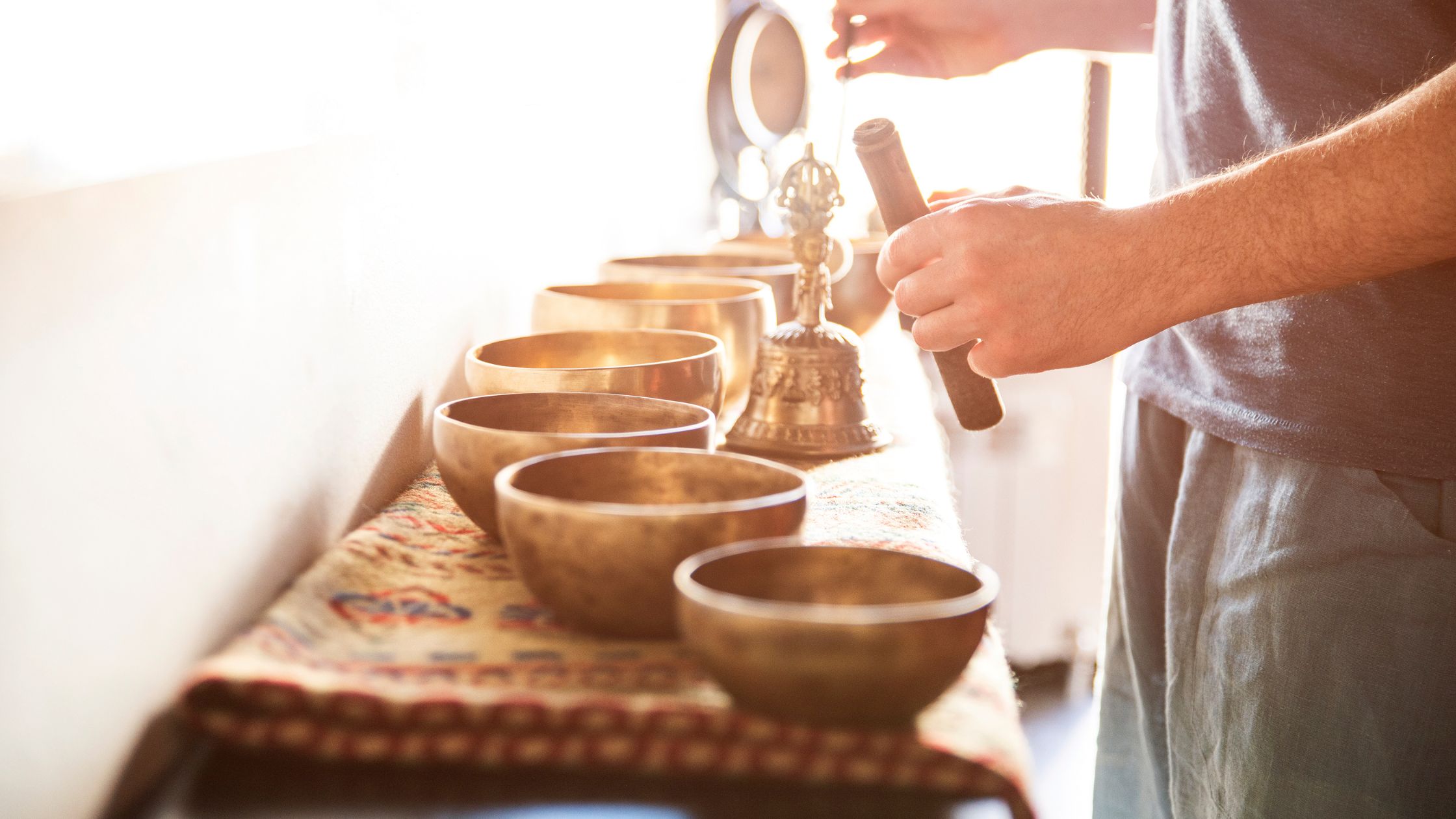 Person playing a row of singing bowls with a mallet, using sound healing techniques in a softly lit space with a patterned cloth.