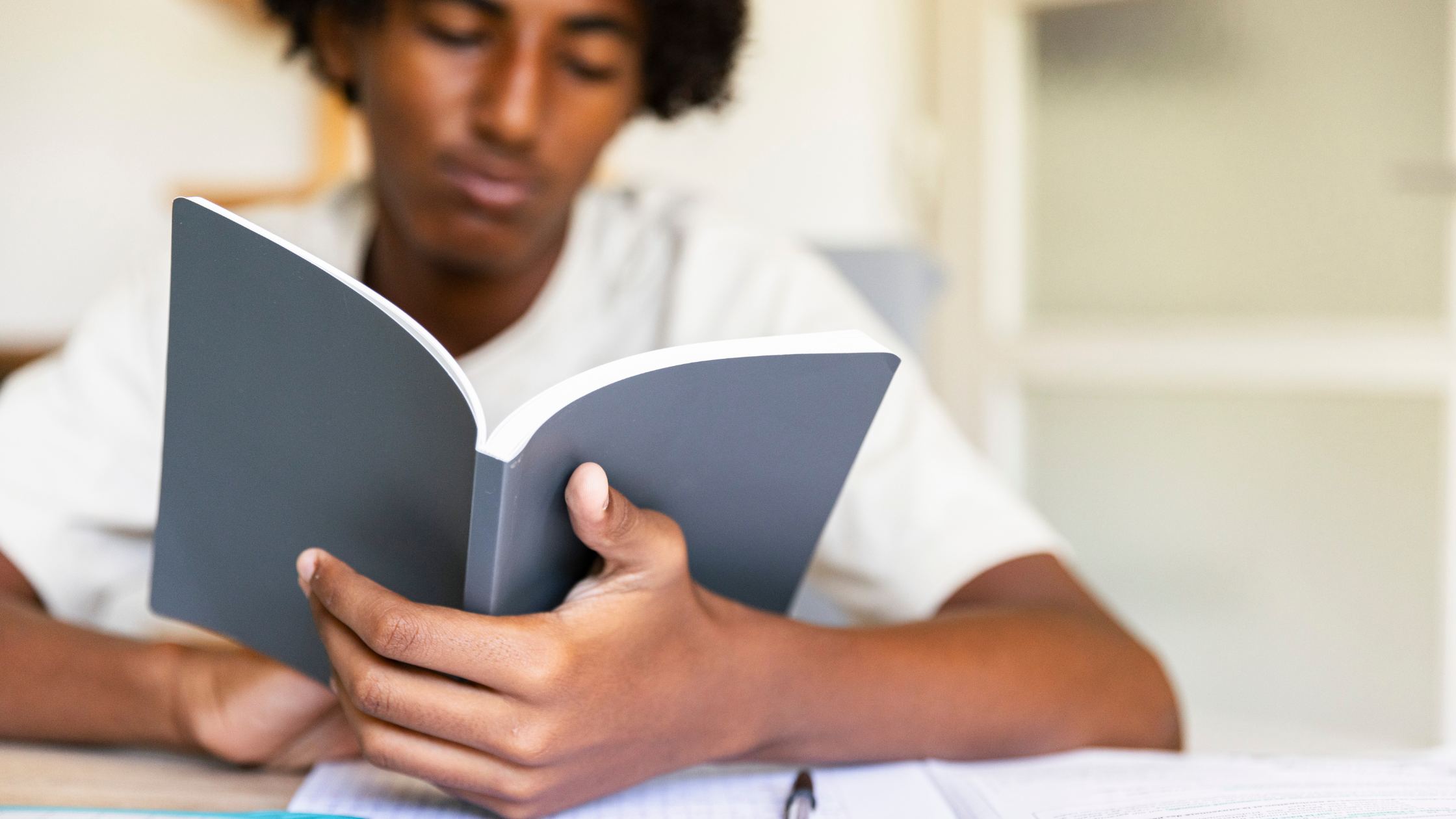 A teenage boy sitting at a desk, focused on reading a book, with notebooks and a pen nearby, symbolizing structured learning and treatment support.