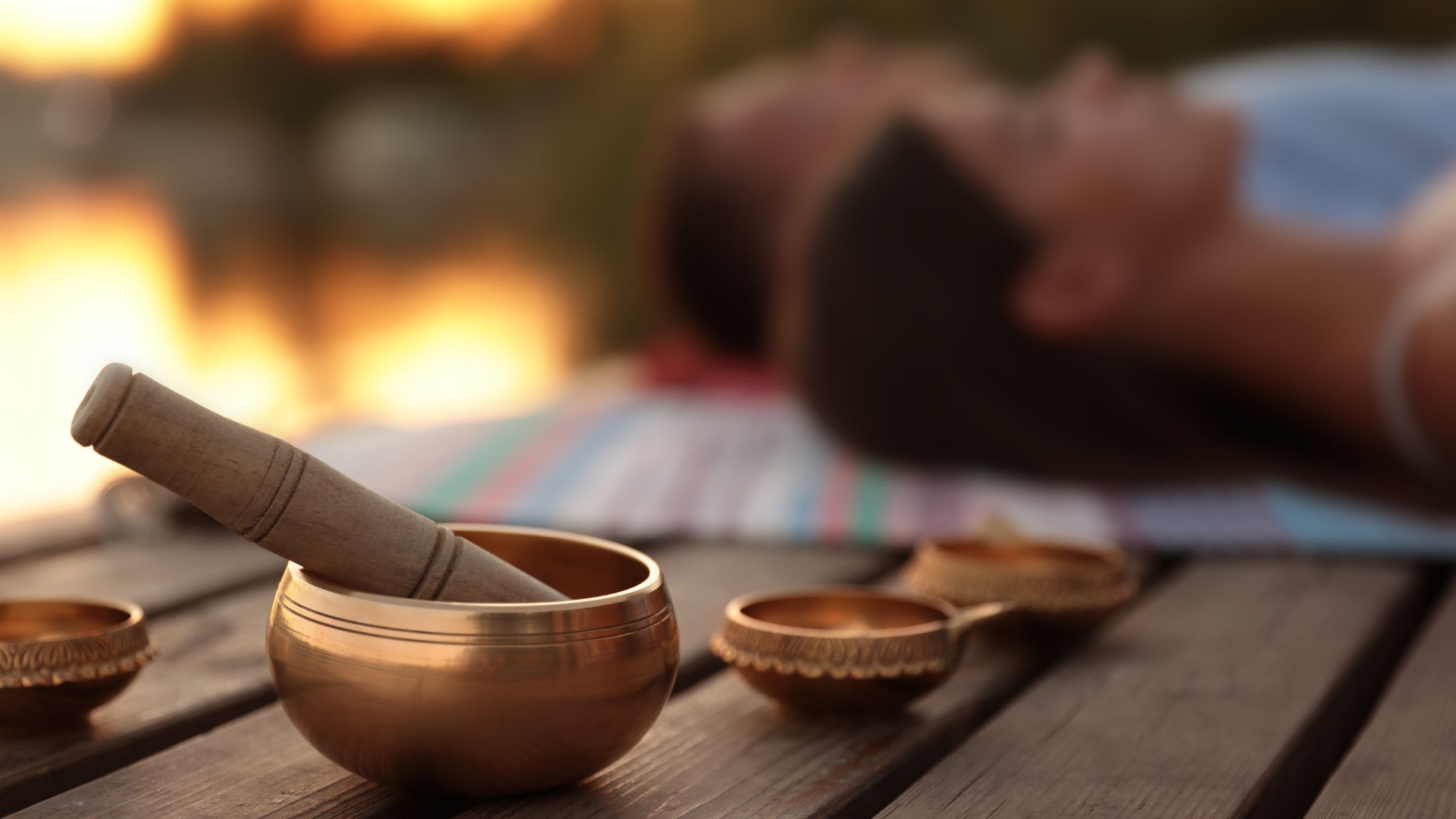Close-up of a Tibetan singing bowl with a wooden striker on a wooden surface, with blurred figures meditating in the background at sunset, symbolizing sound healing therapy.