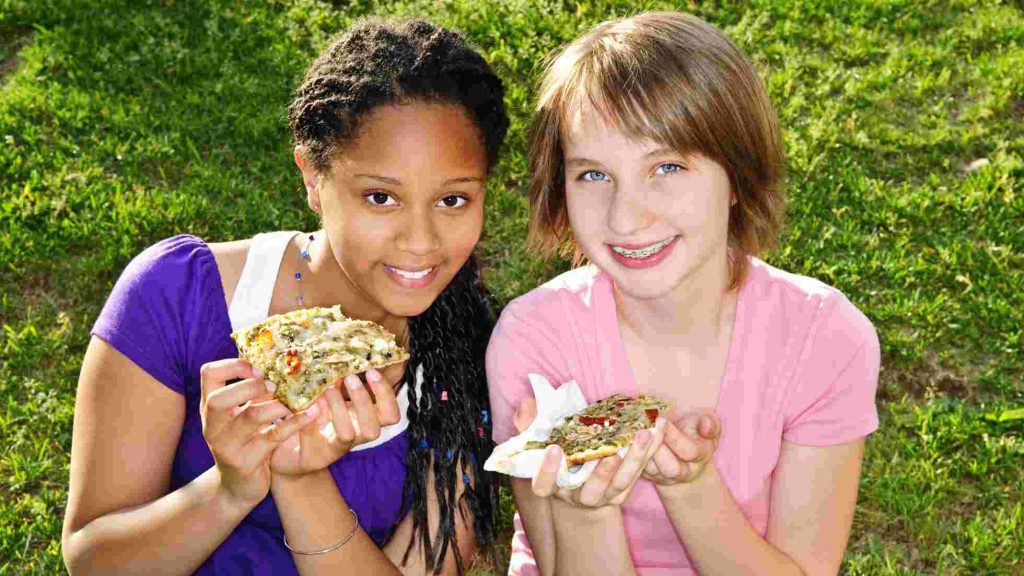 Two smiling teens eating large slices of pizza outdoors, highlighting unhealthy snack choices.