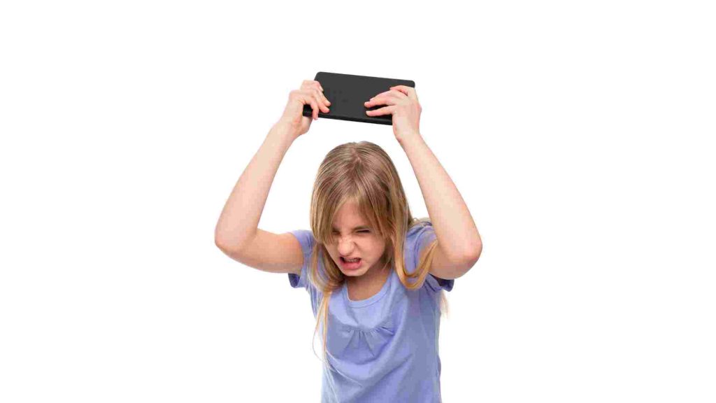 Frustrated teen girl lifts a tablet above her head, teeth clenched and brows furrowed in anger.