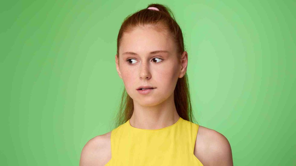 Thoughtful teen in yellow top glancing sideways against a green backdrop, reflecting internal struggle.