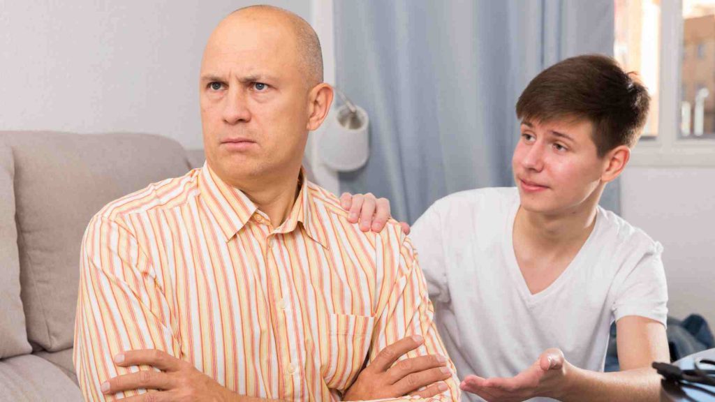 Teen placing hand on worried parent’s shoulder as parent looks away upset, showing tension and disconnect.