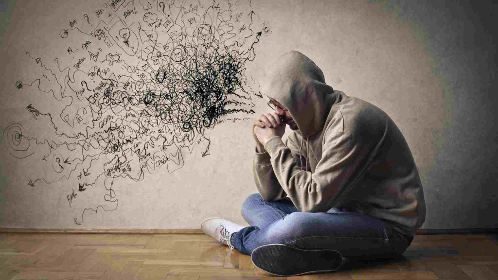Teen in a hoodie sitting on floor, head bowed and hands clasped, surrounded by chaotic thought scribbles.