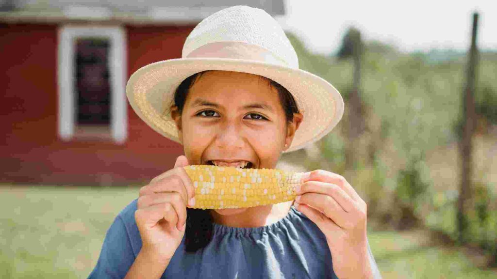 Teen wearing a sun hat happily biting into a corn cob in a sunny outdoor setting.