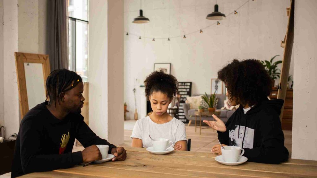 Parents and teen sitting at a table in tense discussion, teen looking down as parents speak.