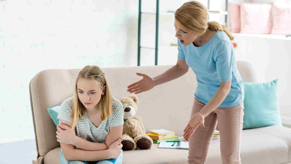 Angry mother leaning forward, gesturing at a withdrawn teenage daughter clutching her arms on the sofa.