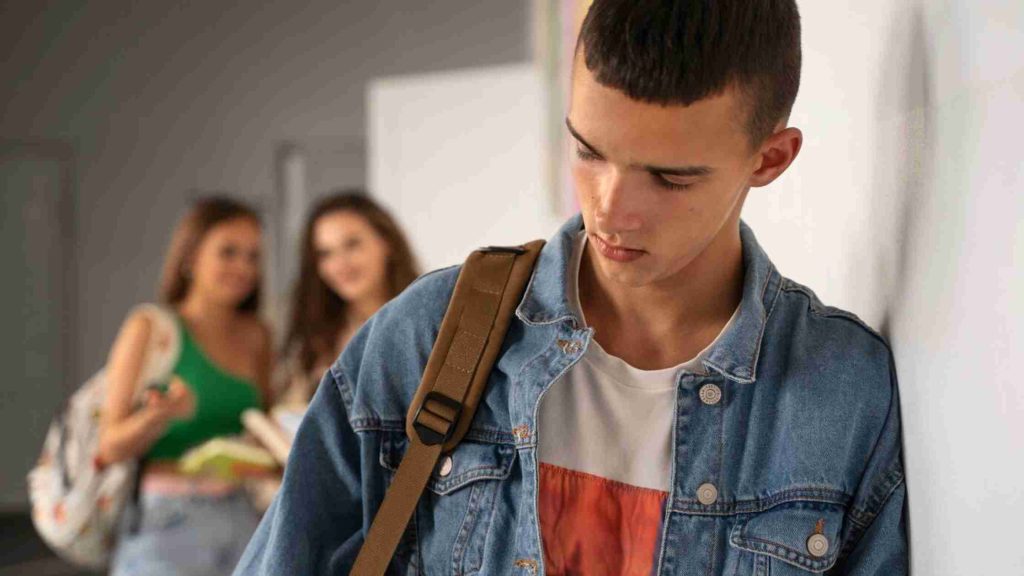 Teen boy leaning against a school hallway wall as peers whisper behind him, symbolizing social isolation and environmental stress.