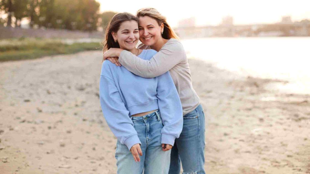 Smiling mother hugs her teenage daughter from behind as they walk together along a sunlit beach.