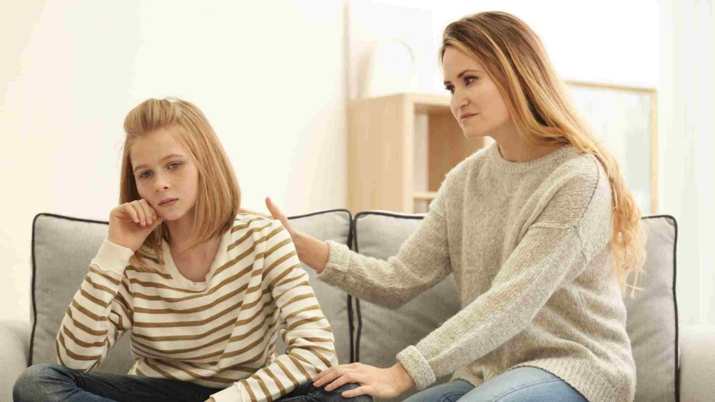 Concerned mother gently rests her hand on her downcast teenage daughter’s shoulder on the living room couch.