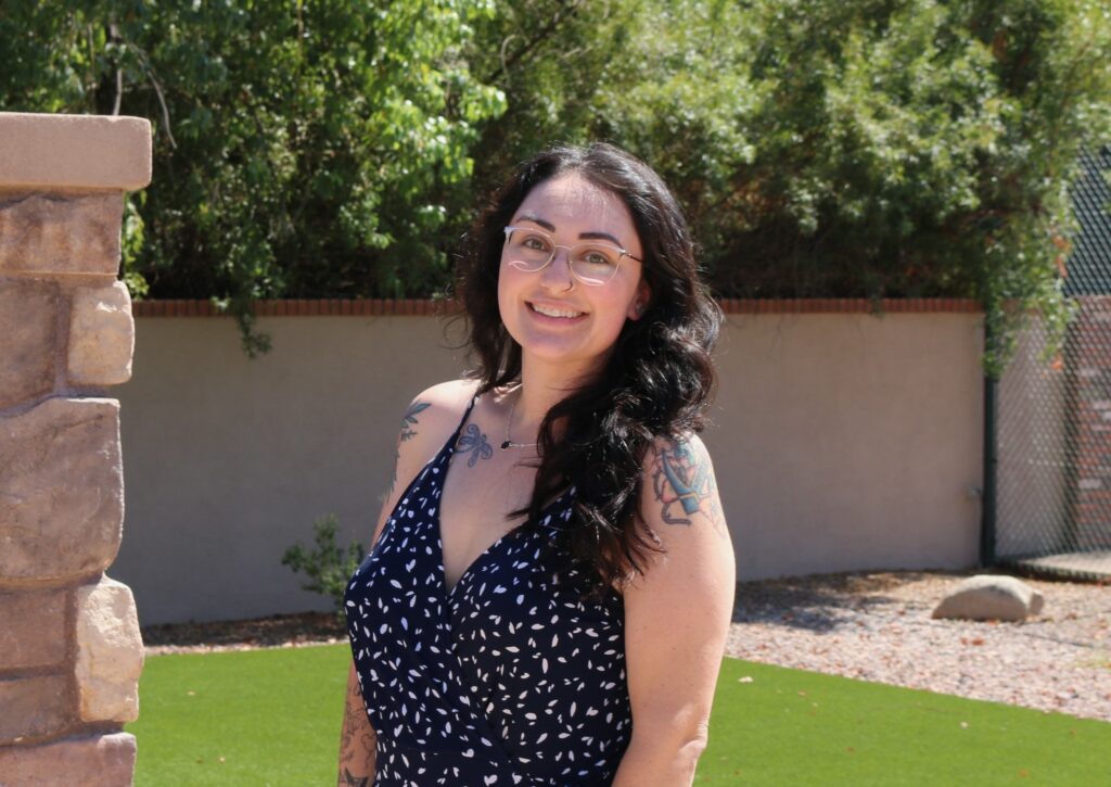 Smiling woman with long dark hair and round glasses, wearing a navy polka-dot dress, standing beside a stone pillar outdoors.