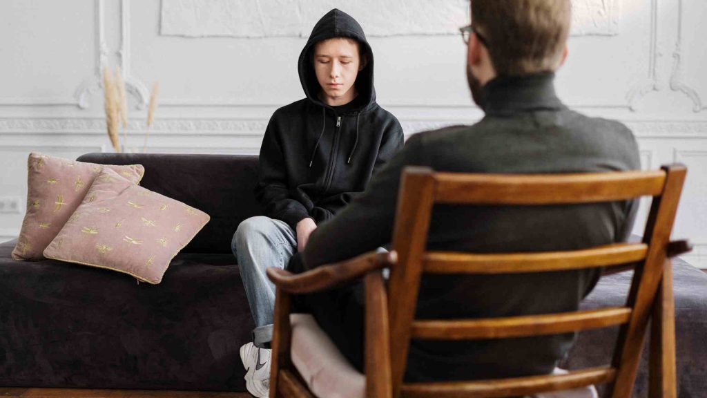Teen boy in hoodie talking with therapist in office, representing therapy as a key factor in choosing the right boys’ home.