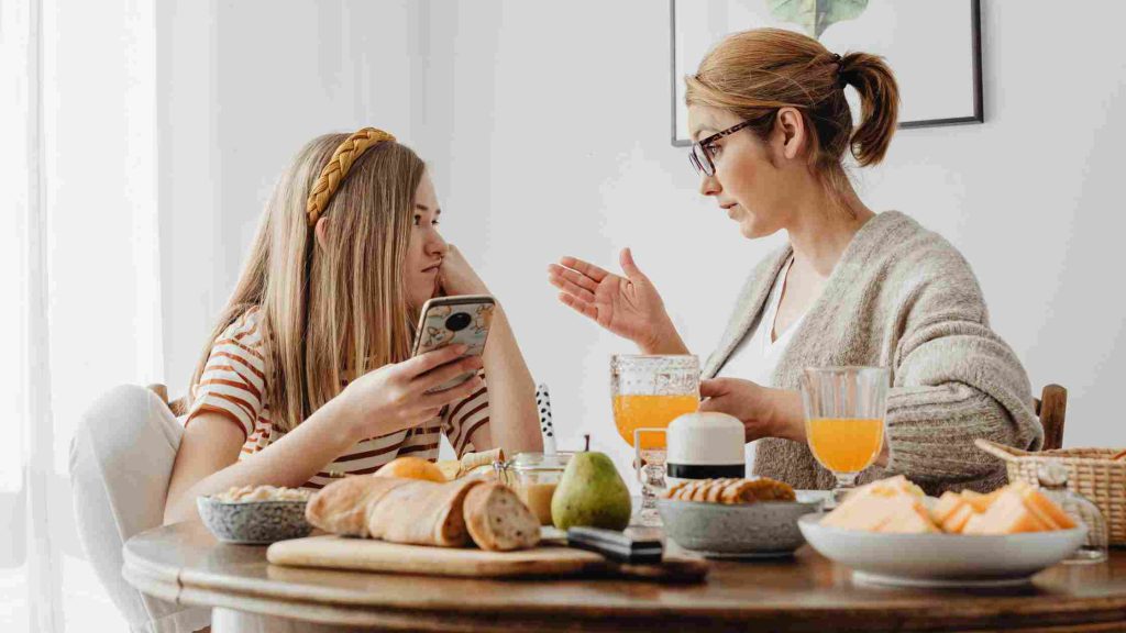 Mother calmly talking to her teenage daughter using a phone during breakfast, showing how to handle teen disobedience.