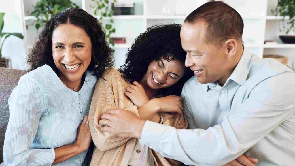 Parents sit close and embrace their teenage child on a couch, smiling & offering comfort, reassurance, and emotional support.