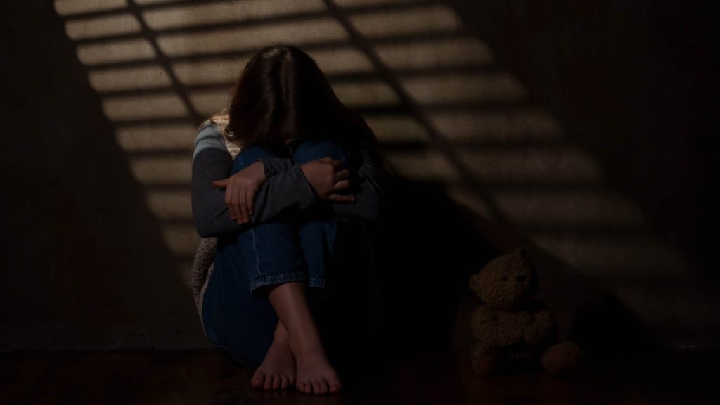 Teen sits alone on the floor in a dark room, hugging knees with head down, showing isolation, sadness, & emotional distress.