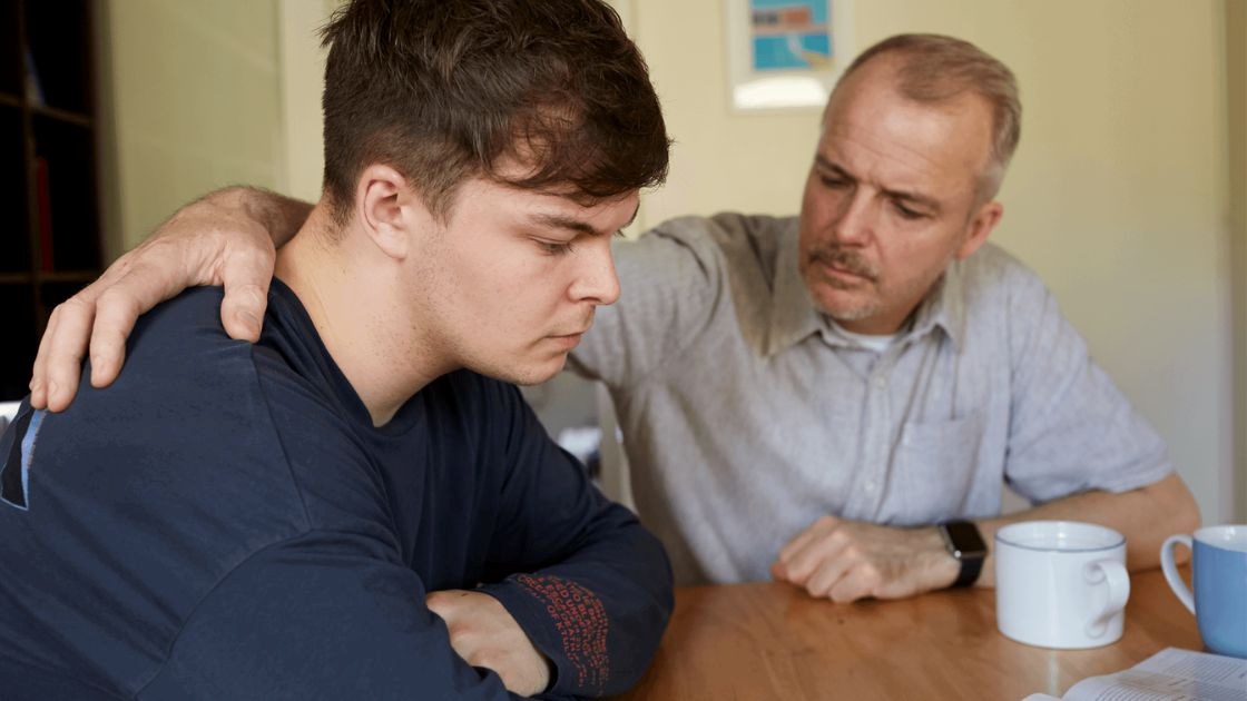 Father comforting distressed teen at table, reflecting supportive conversations about intrusive thoughts and OCD struggles.